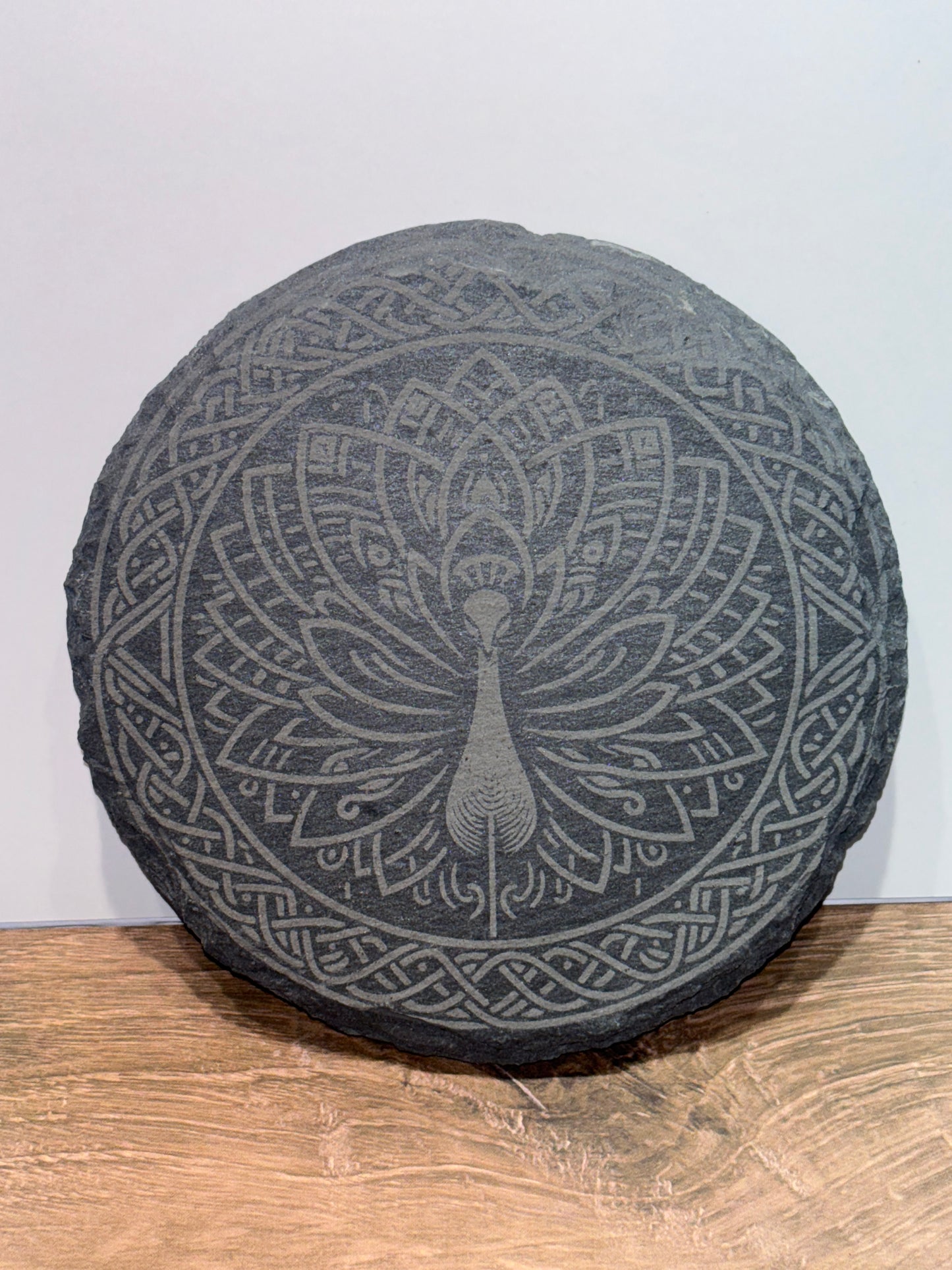 Round natural slate plaque with an intricate Celtic-style peacock mandala engraving, displayed on a wooden surface.