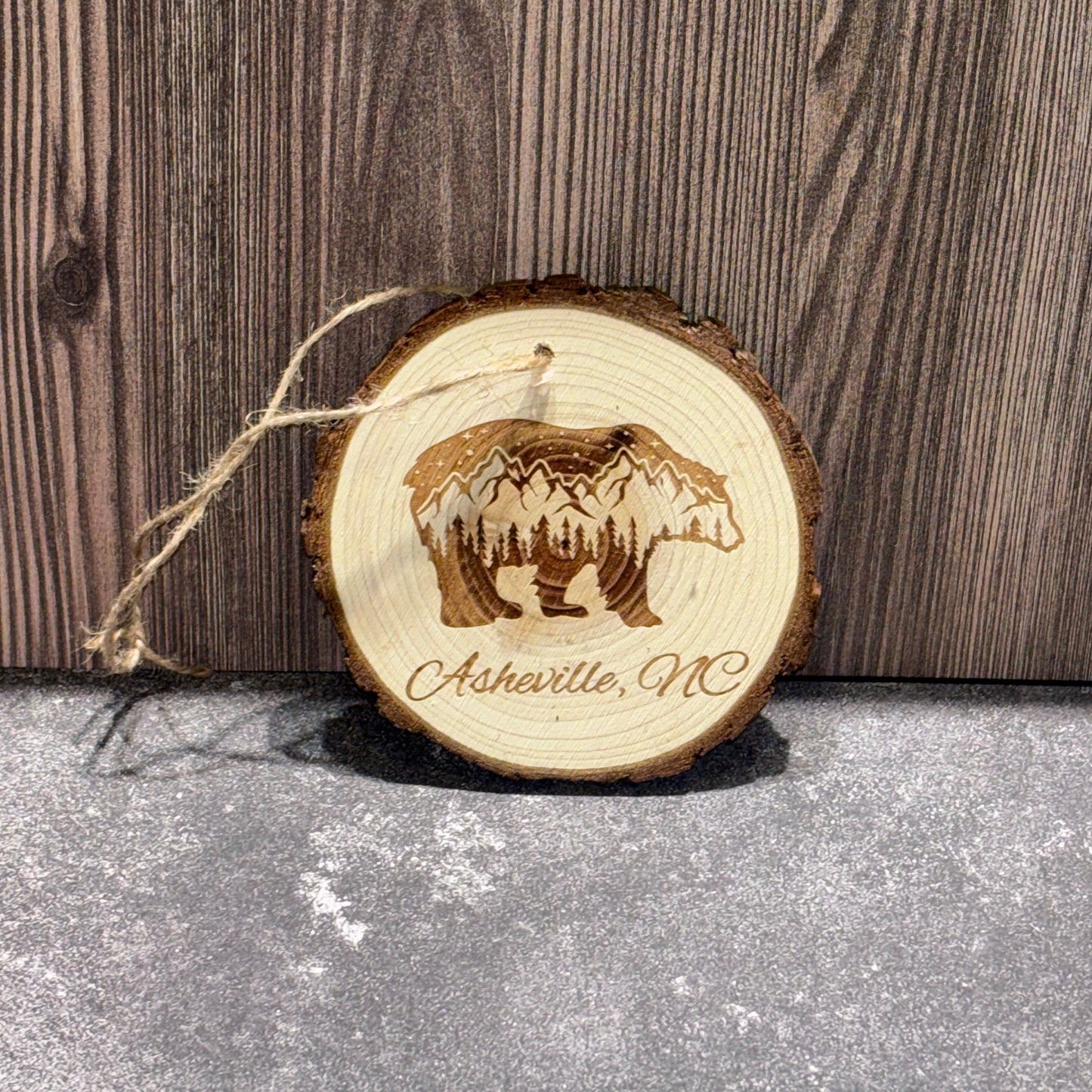 4 inch natural wood slice ornament engraved with a bear silhouette containing mountain and forest scenery and the words Asheville, NC.