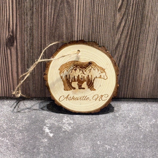 4 inch natural wood slice ornament engraved with a bear silhouette containing mountain and forest scenery and the words Asheville, NC.