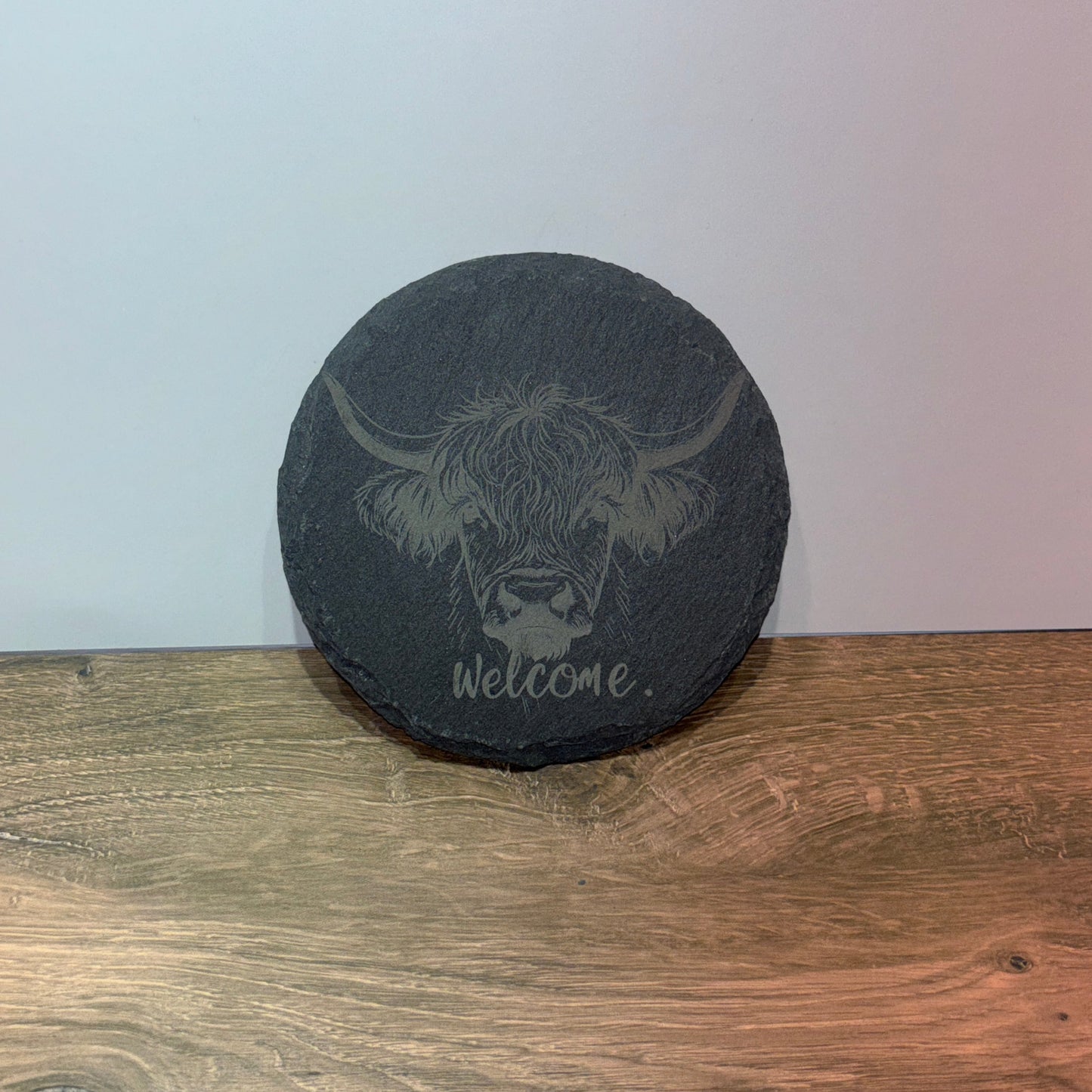Round 4 inch black slate coaster engraved with a Highland cow face and the word “Welcome.”