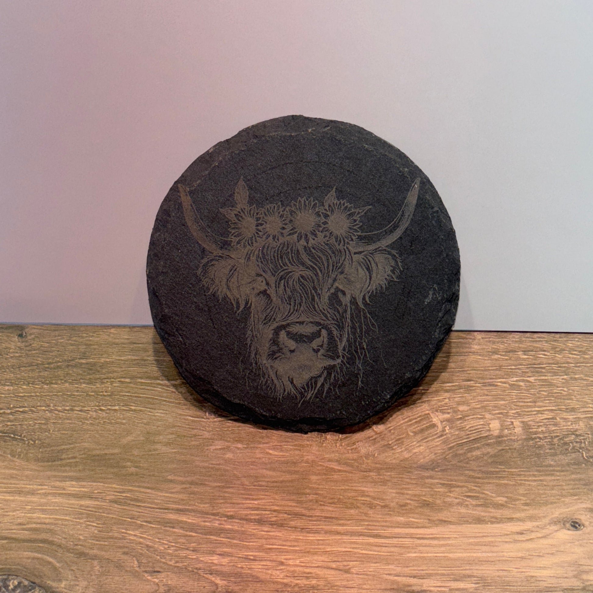 Round natural slate plaque with a laser-engraved Highland cow wearing a sunflower crown, displayed on a wooden surface.
