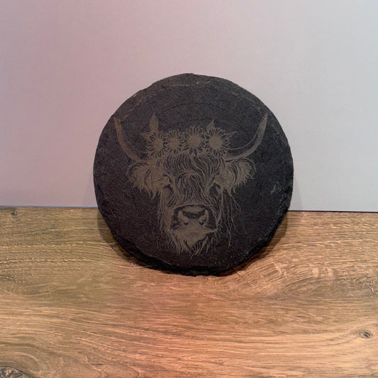 Round natural slate plaque with a laser-engraved Highland cow wearing a sunflower crown, displayed on a wooden surface.