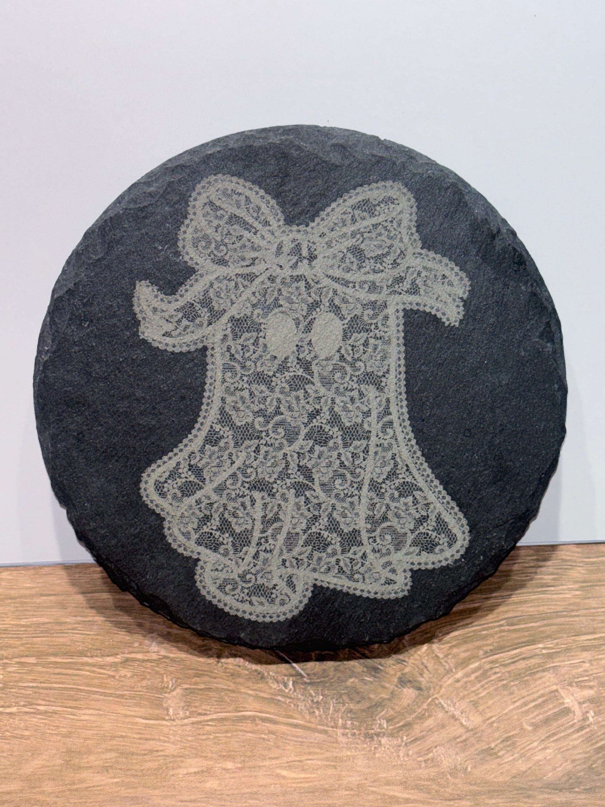 Round natural slate plaque featuring a laser-engraved lace-style ghost with a bow, displayed on a wooden surface.