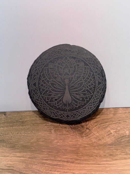 Round natural slate plaque with an intricate Celtic-style peacock mandala engraving, displayed on a wooden surface.