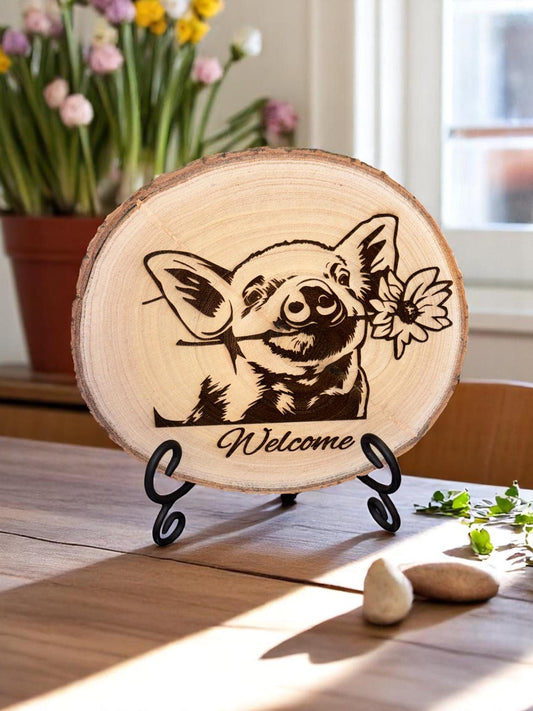 Wooden plaque with a pig illustration and 'Welcome' text on a stand, with flowers in the background.